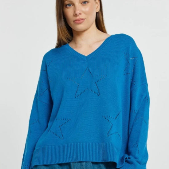 A woman wearing a bright blue knit sweater with star-shaped cutout details on the front, paired with blue jeans, against a plain light gray studio background.