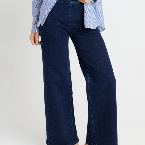 A person wearing dark blue wide-leg denim jeans, a light blue and white striped shirt, and beige slip-on shoes, shown from the waist down against a light gray studio background.