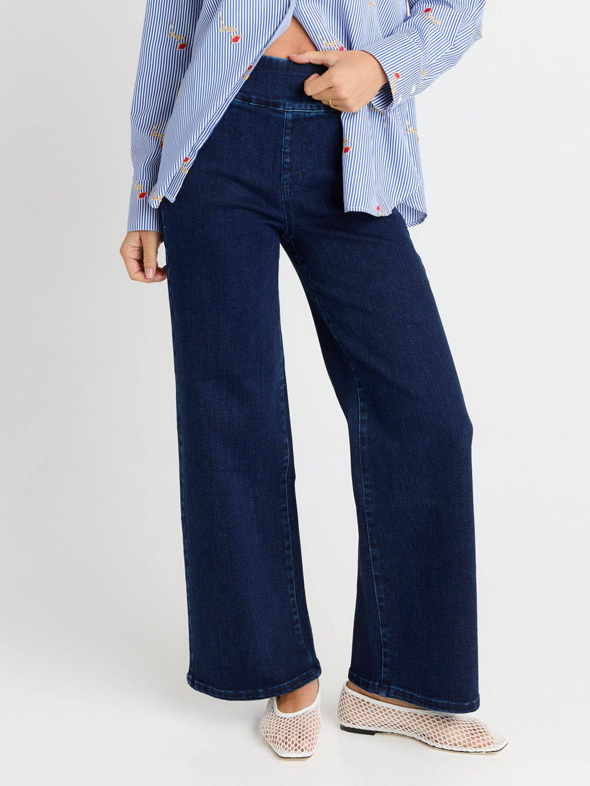 A person wearing dark blue wide-leg denim jeans, a light blue and white striped shirt, and beige slip-on shoes, shown from the waist down against a light gray studio background.