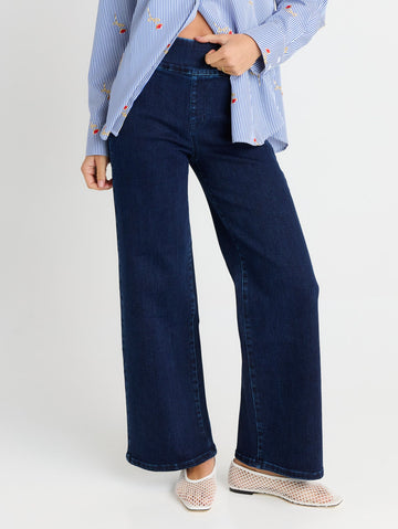 A person wearing dark blue wide-leg denim jeans, a light blue and white striped shirt, and beige slip-on shoes, shown from the waist down against a light gray studio background.