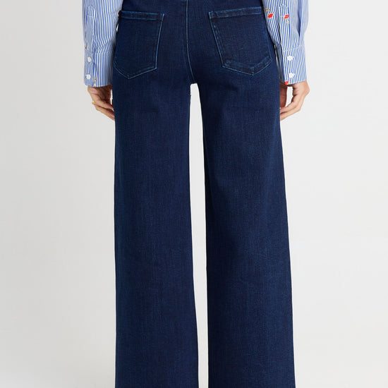 Back view of a person wearing dark blue wide-leg denim jeans with a light blue and white striped shirt and white shoes, against a light gray studio background.