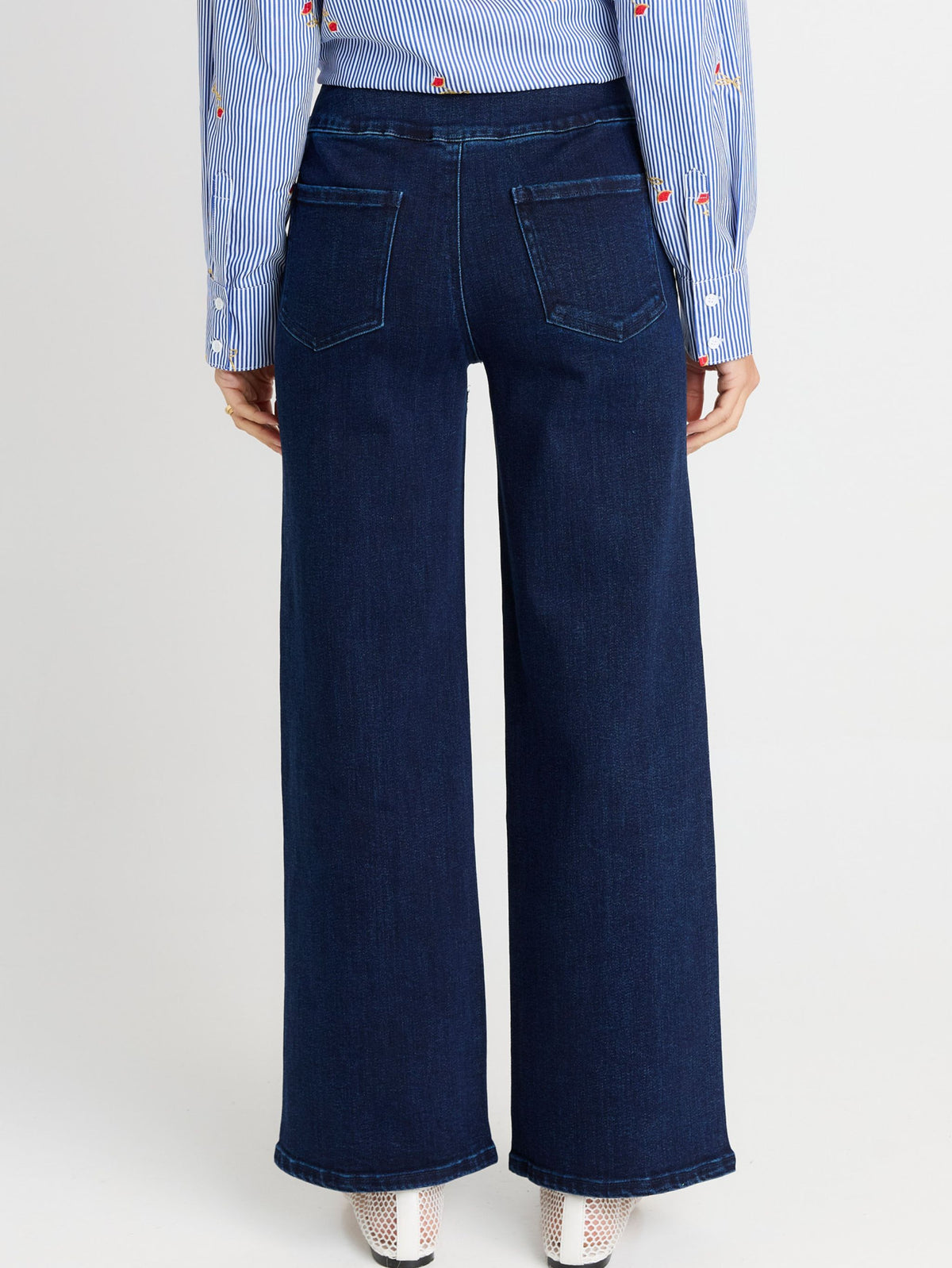 Back view of a person wearing dark blue wide-leg denim jeans with a light blue and white striped shirt and white shoes, against a light gray studio background.