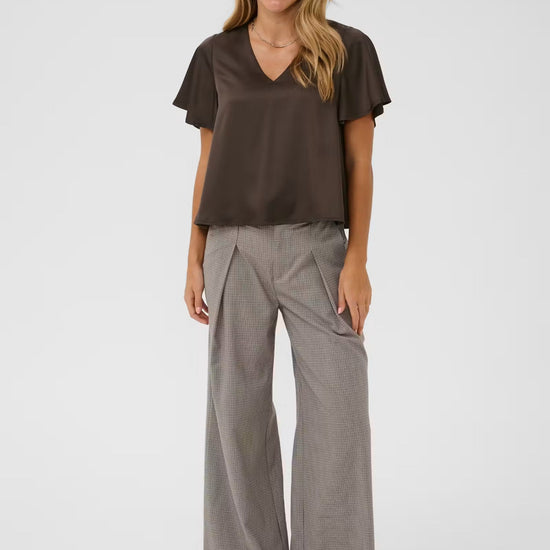 A woman modeling a dark brown short-sleeve V-neck top and light gray wide-leg trousers against a plain light gray studio background.