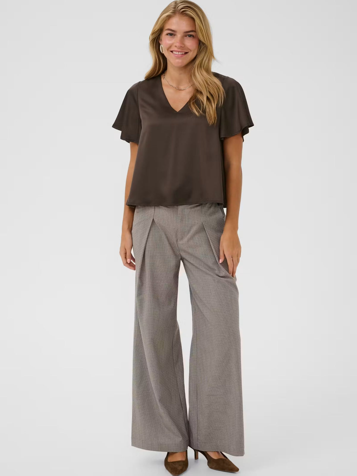 A woman modeling a dark brown short-sleeve V-neck top and light gray wide-leg trousers against a plain light gray studio background.