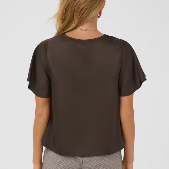 A woman viewed from the back wearing a dark brown short-sleeve blouse with flutter sleeves and gray patterned trousers, against a light gray studio background.