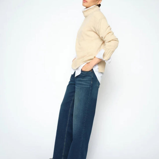 A model wearing a beige ribbed turtleneck sweater, a white collared shirt, and wide-leg dark blue jeans, standing against a plain white studio background.