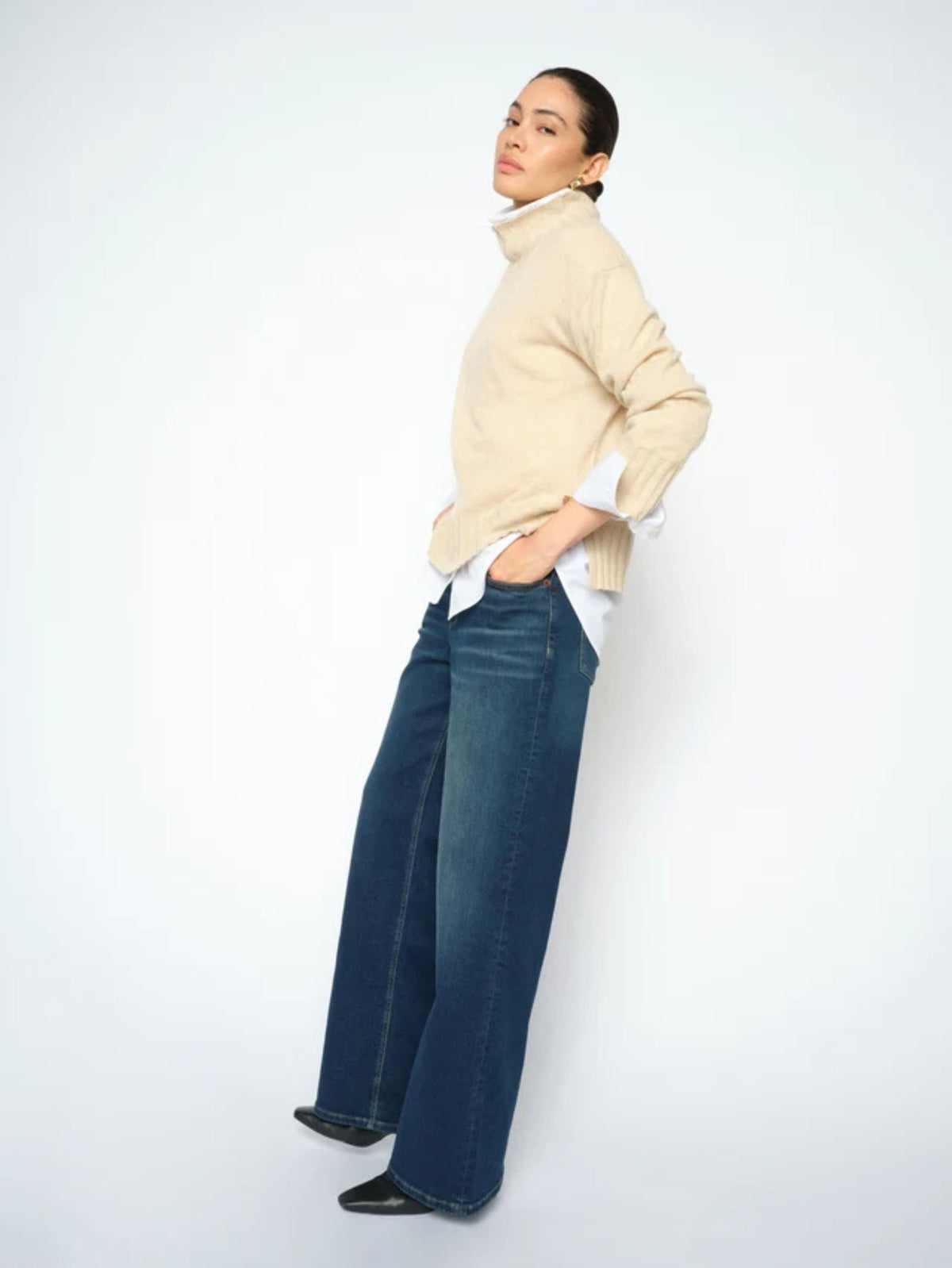 A model wearing a beige ribbed turtleneck sweater, a white collared shirt, and wide-leg dark blue jeans, standing against a plain white studio background.