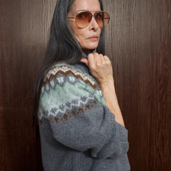 A woman with long gray hair and aviator sunglasses poses against a dark wood-paneled wall, wearing a gray knit sweater with a green, brown, and white Fair Isle patterned yoke.