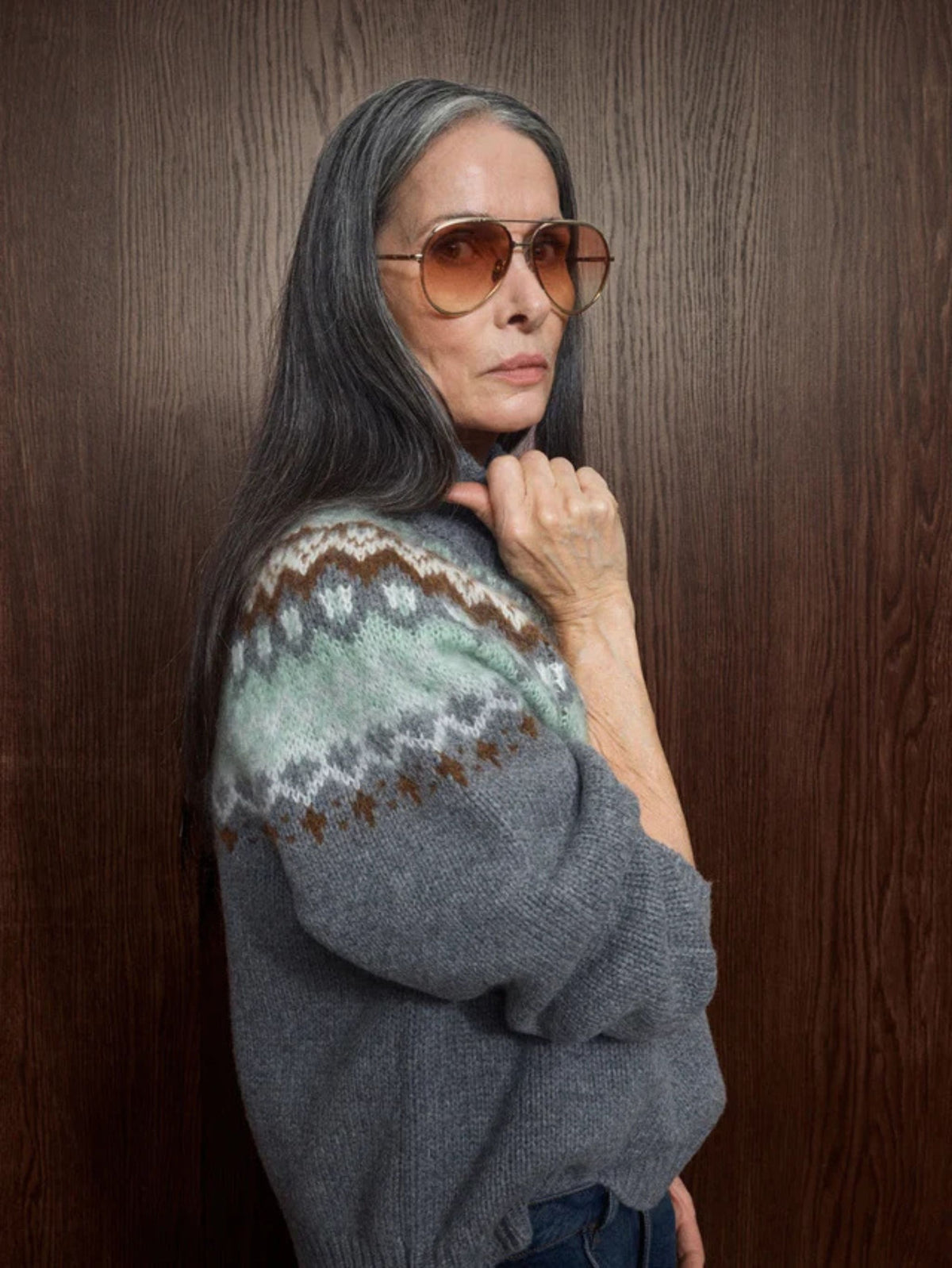A woman with long gray hair and aviator sunglasses poses against a dark wood-paneled wall, wearing a gray knit sweater with a green, brown, and white Fair Isle patterned yoke.