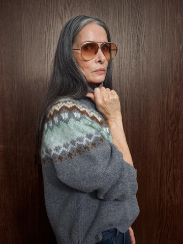 A woman with long gray hair and aviator sunglasses poses against a dark wood-paneled wall, wearing a gray knit sweater with a green, brown, and white Fair Isle patterned yoke.