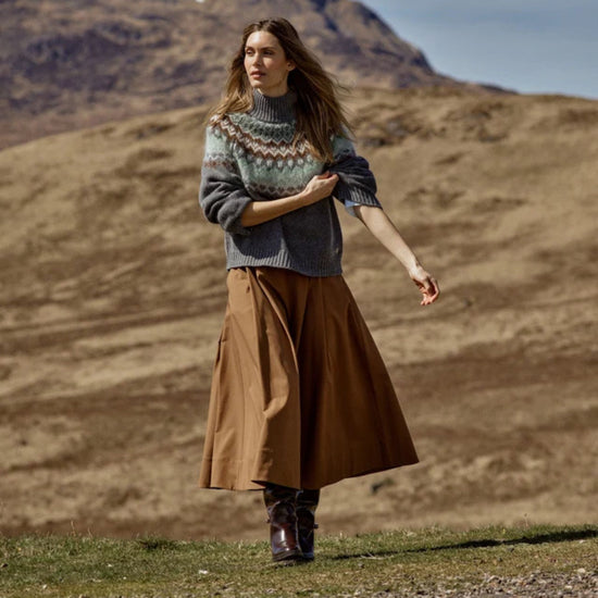 Woman in a patterned sweater and brown skirt standing in a mountainous landscape.