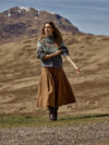 Woman in a patterned sweater and brown skirt standing in a mountainous landscape.