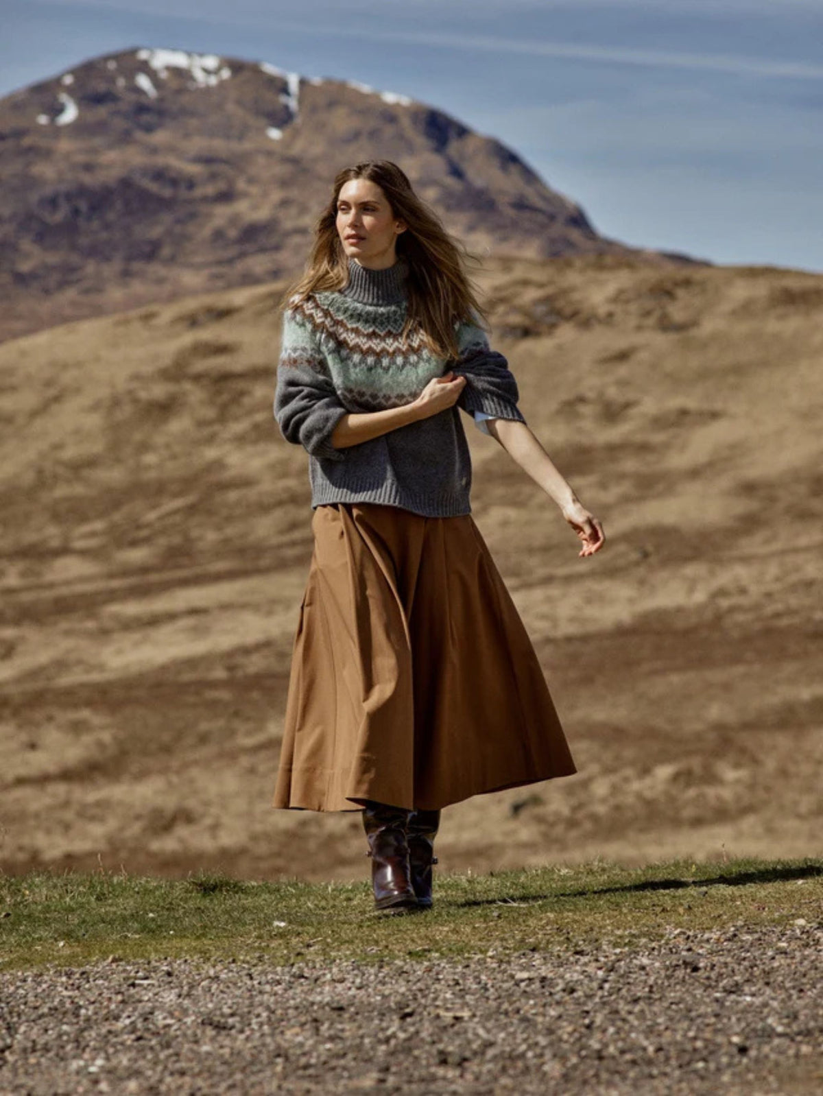 Woman in a patterned sweater and brown skirt standing in a mountainous landscape.