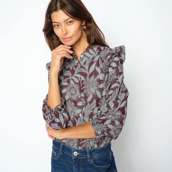 A woman modeling a long-sleeve blouse with a gray and burgundy floral pattern and ruffled shoulders, paired with a dark blue denim jeans, against a plain white studio background.