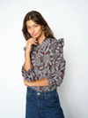 A woman modeling a long-sleeve blouse with a gray and burgundy floral pattern and ruffled shoulders, paired with a dark blue denim jeans, against a plain white studio background.