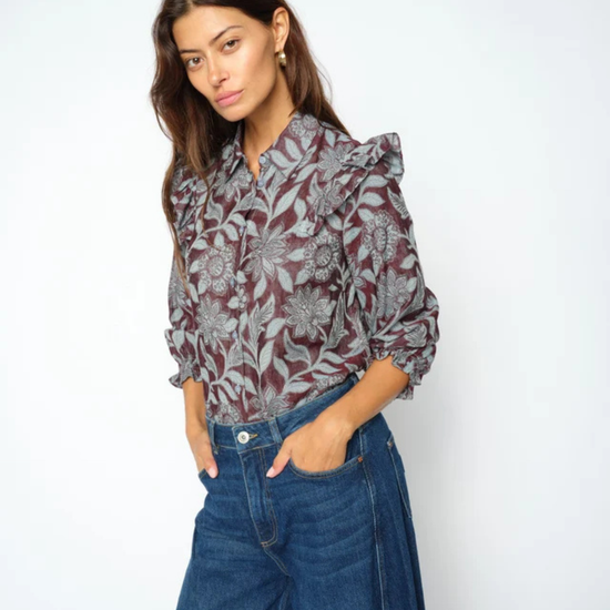 A woman modeling a long-sleeve button-up blouse with a gray and burgundy floral pattern, paired with high-waisted blue jeans, against a plain white studio background.