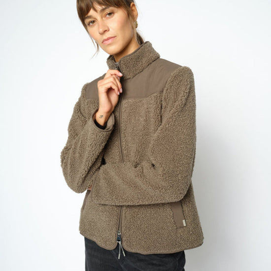 A woman wearing a olive sherpa fleece jacket with a high collar and front zipper, paired with dark pants, photographed against a light gray studio background.