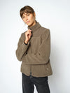 A woman wearing a olive sherpa fleece jacket with a high collar and front zipper, paired with dark pants, photographed against a light gray studio background.