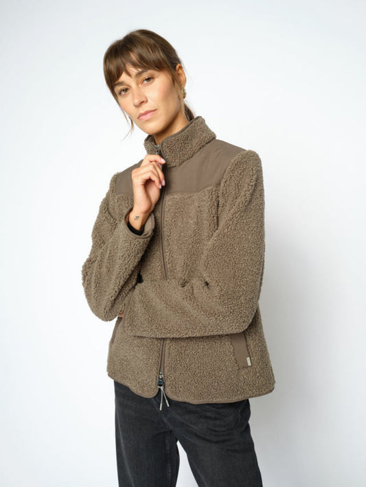 A woman wearing a olive sherpa fleece jacket with a high collar and front zipper, paired with dark pants, photographed against a light gray studio background.