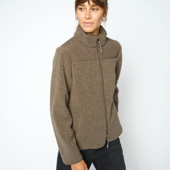 A woman modeling a olive fleece jacket with a mock neck and zippered front, paired with dark jeans, against a plain light gray background.