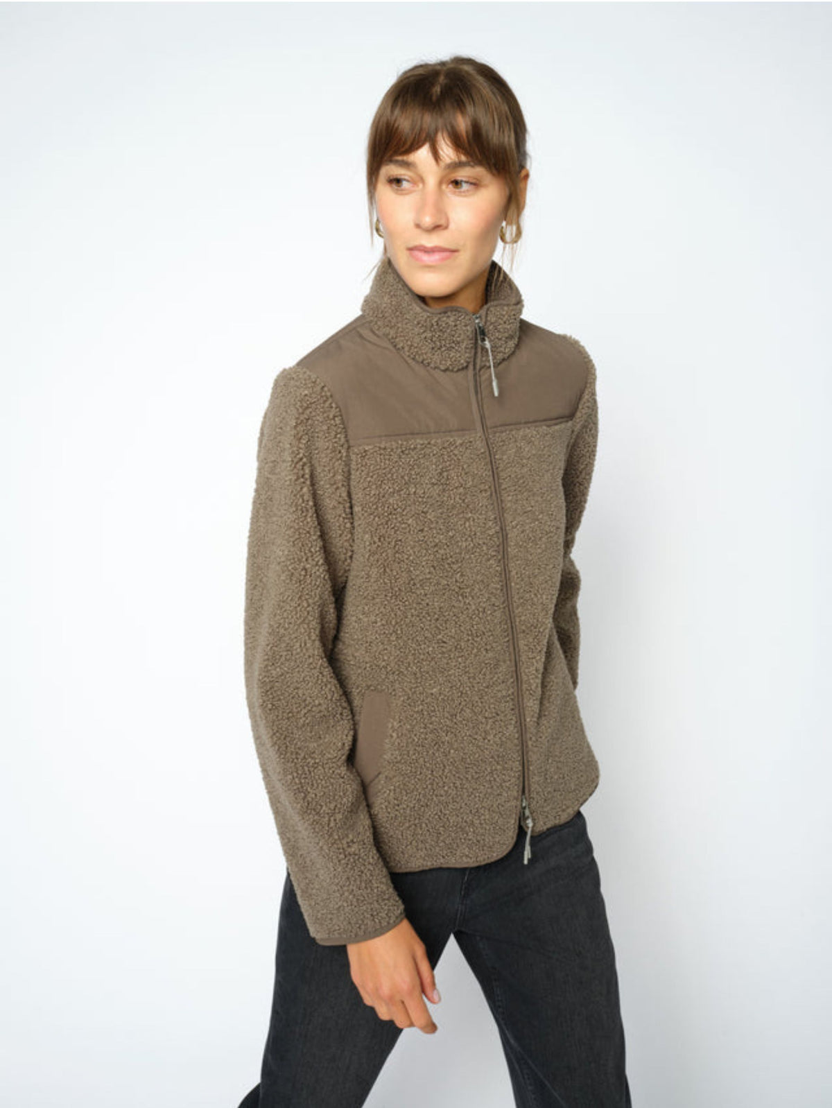 A woman modeling a olive fleece jacket with a mock neck and zippered front, paired with dark jeans, against a plain light gray background.