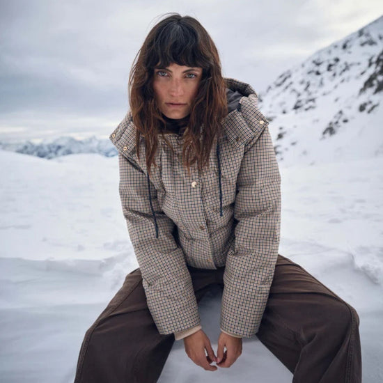 A model wearing a beige and brown checkered puffer jacket and dark brown pants, sitting in a snowy mountain landscape under an overcast sky.