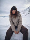 A model wearing a beige and brown checkered puffer jacket and dark brown pants, sitting in a snowy mountain landscape under an overcast sky.