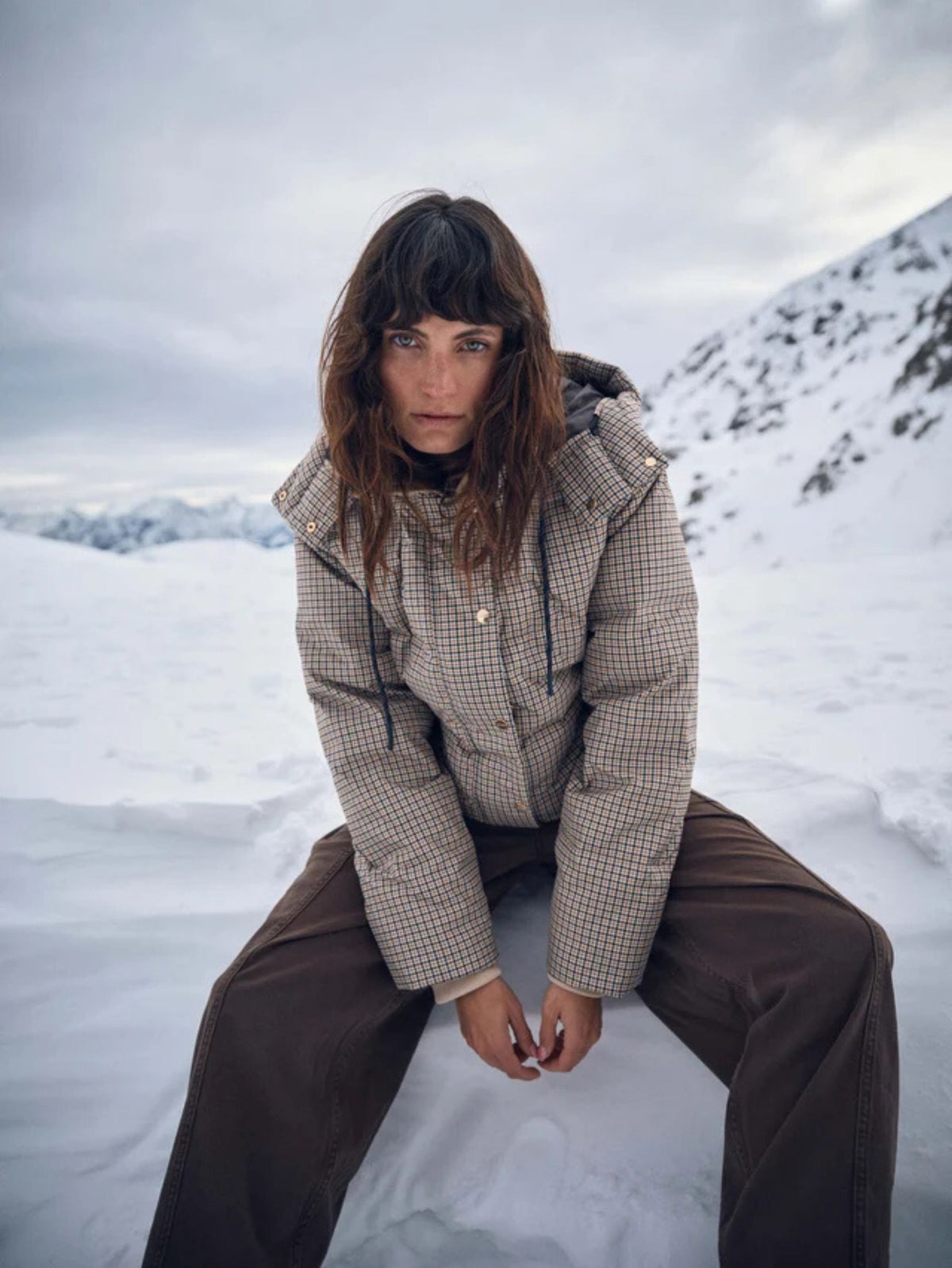 A model wearing a beige and brown checkered puffer jacket and dark brown pants, sitting in a snowy mountain landscape under an overcast sky.