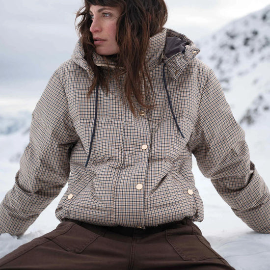 A woman wearing a beige and brown checkered puffer jacket and dark brown pants, sitting outdoors in a snowy mountain setting.