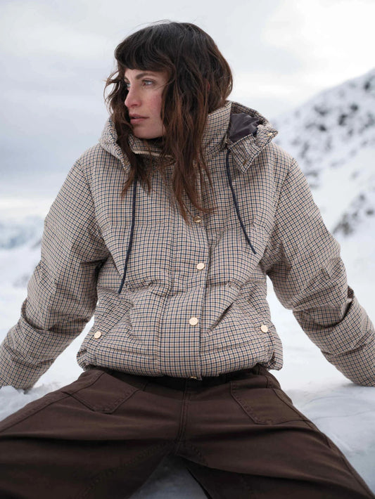 A woman wearing a beige and brown checkered puffer jacket and dark brown pants, sitting outdoors in a snowy mountain setting.
