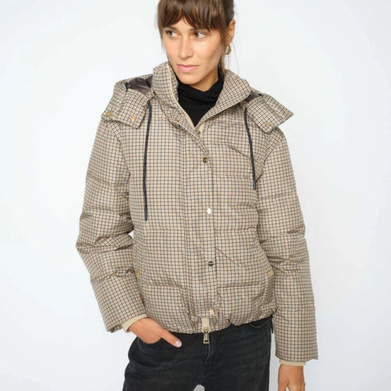 A woman wearing a beige and brown checkered puffer jacket with a high collar and black turtleneck underneath, paired with black jeans, against a plain light gray background.