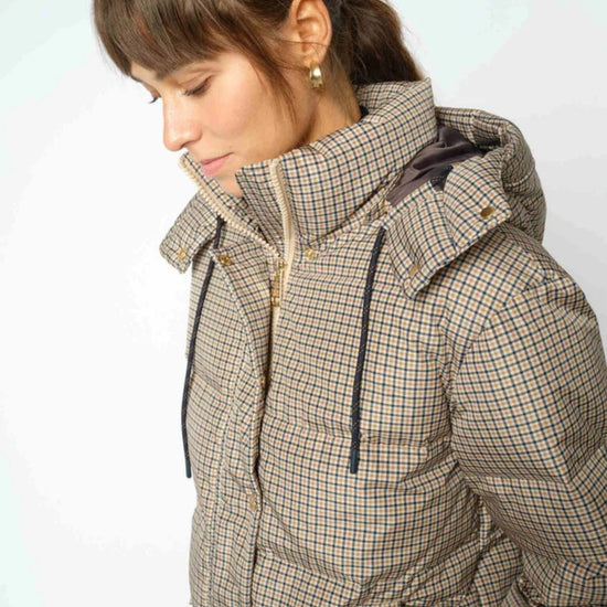 A woman wearing a beige and brown checkered puffer jacket with a high collar and hood, shown in a close-up profile view against a light gray background.