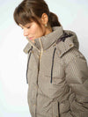 A woman wearing a beige and brown checkered puffer jacket with a high collar and hood, shown in a close-up profile view against a light gray background.