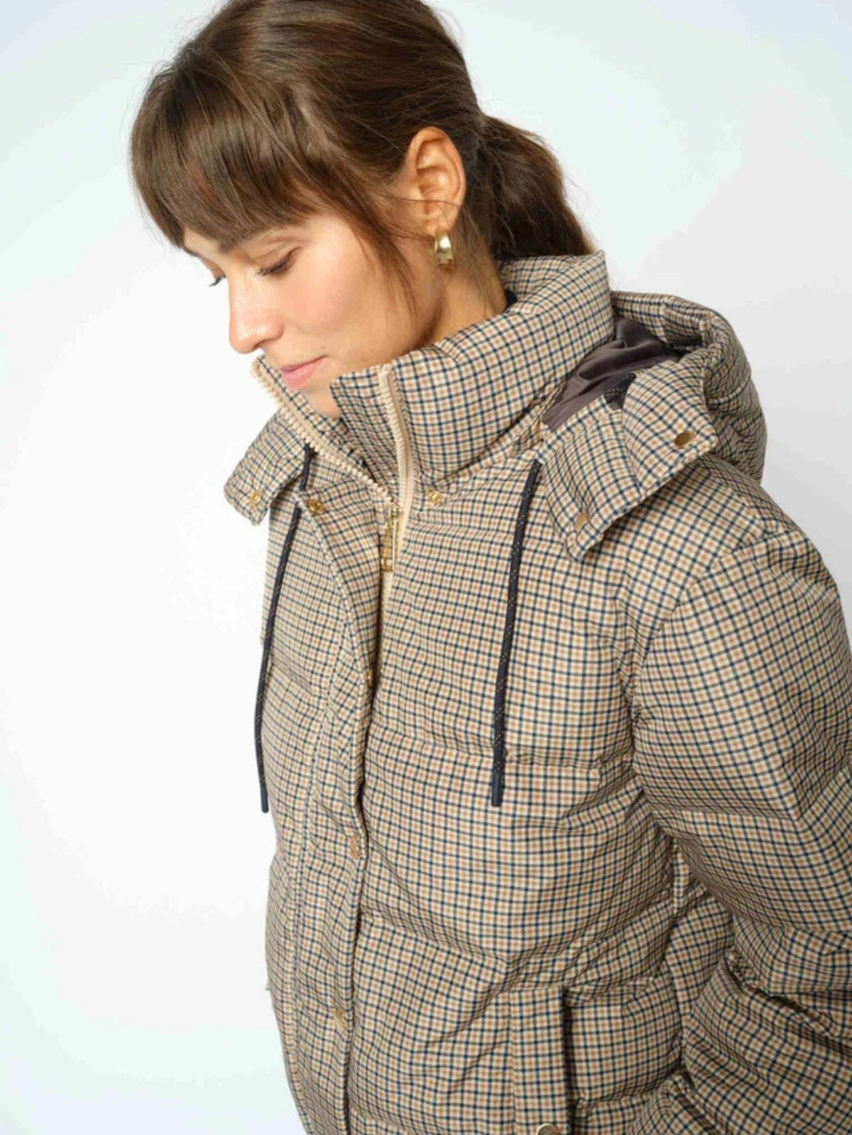 A woman wearing a beige and brown checkered puffer jacket with a high collar and hood, shown in a close-up profile view against a light gray background.