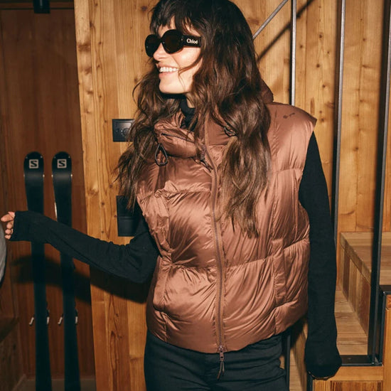 A woman wearing a shiny bronze-brown puffer vest over a black long-sleeve top and black pants, standing in a wooden interior near ski equipment. 