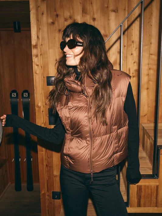 A woman wearing a shiny bronze-brown puffer vest over a black long-sleeve top and black pants, standing in a wooden interior near ski equipment. 