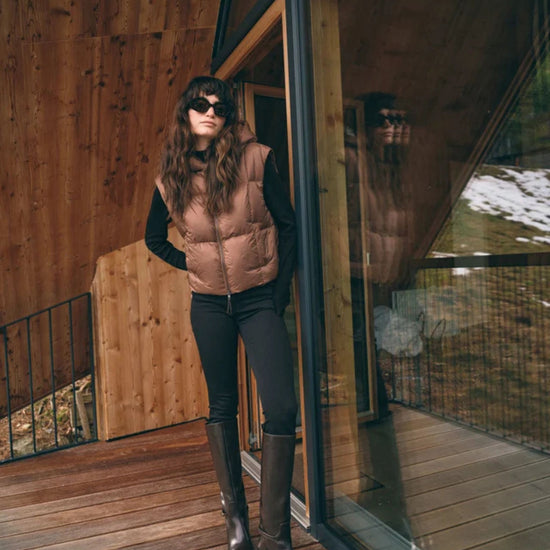 A woman stands on a wooden deck outside a modern cabin wearing a tan quilted puffer vest over a black long-sleeve top, black pants, and dark brown knee-high boots.