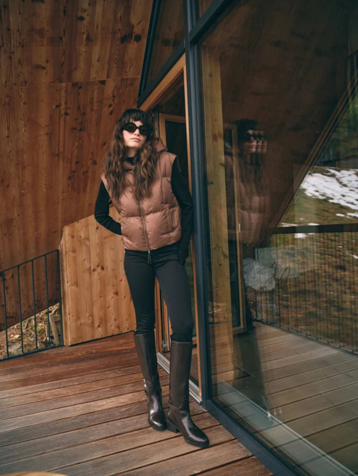 A woman stands on a wooden deck outside a modern cabin wearing a tan quilted puffer vest over a black long-sleeve top, black pants, and dark brown knee-high boots.