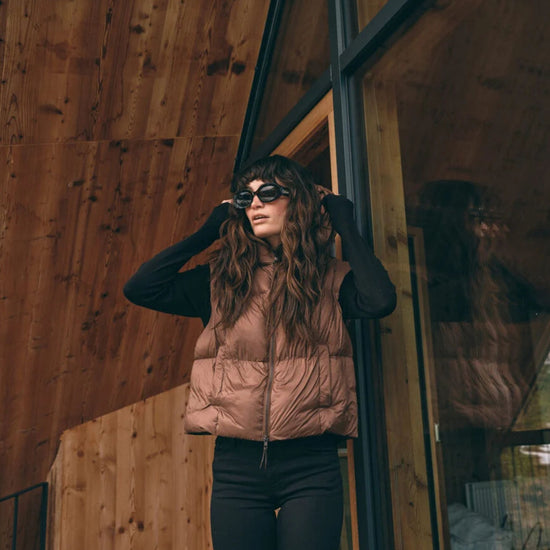 A woman wearing a light brown puffer vest over a black long-sleeve top and black pants, standing in front of a wooden wall and glass window of a modern building.