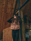 A woman wearing a light brown puffer vest over a black long-sleeve top and black pants, standing in front of a wooden wall and glass window of a modern building.