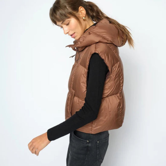 A woman wearing a brown puffer vest over a black long-sleeve top and dark jeans, standing against a light gray studio background.
