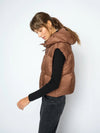A woman wearing a brown puffer vest over a black long-sleeve top and dark jeans, standing against a light gray studio background.