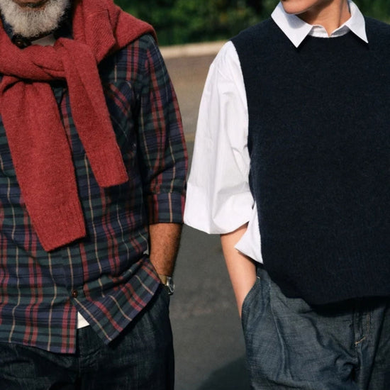 Two people standing outdoors, one wearing a red scarf and plaid shirt, the other in a black vest and white shirt.