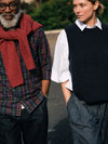 Two people standing outdoors, one wearing a red scarf and plaid shirt, the other in a black vest and white shirt.