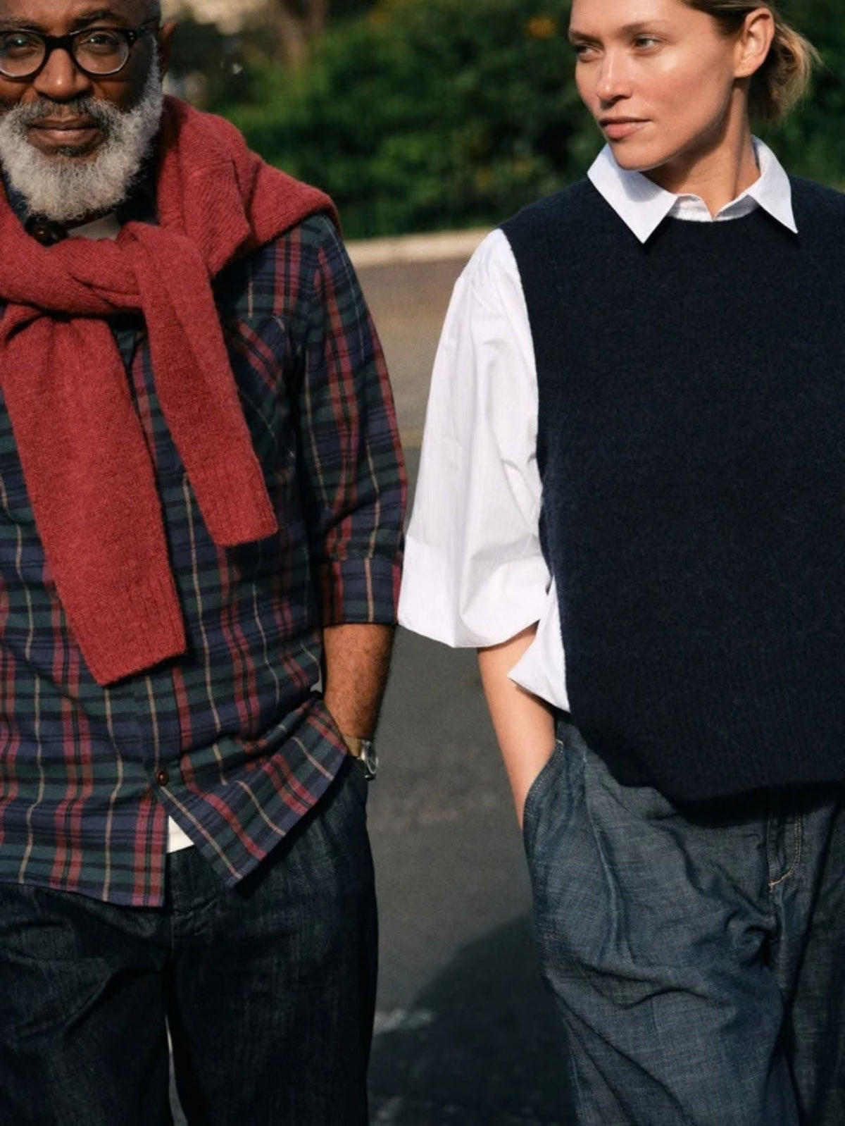 Two people standing outdoors, one wearing a red scarf and plaid shirt, the other in a black vest and white shirt.