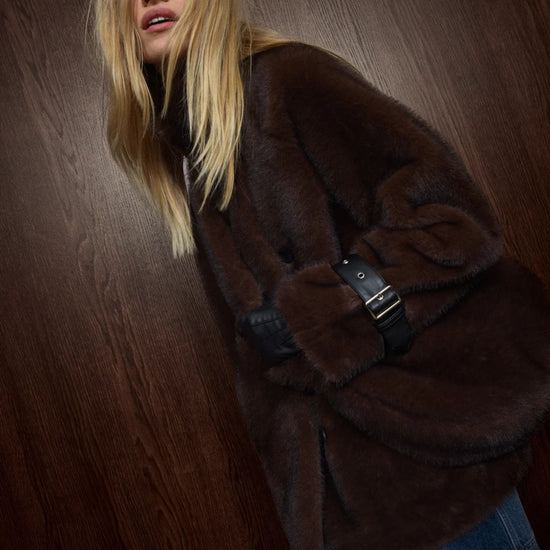 A woman wearing a dark brown plush faux fur coat with a black belt detail, posed against a dark wood-paneled background.