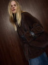 A woman wearing a dark brown plush faux fur coat with a black belt detail, posed against a dark wood-paneled background.
