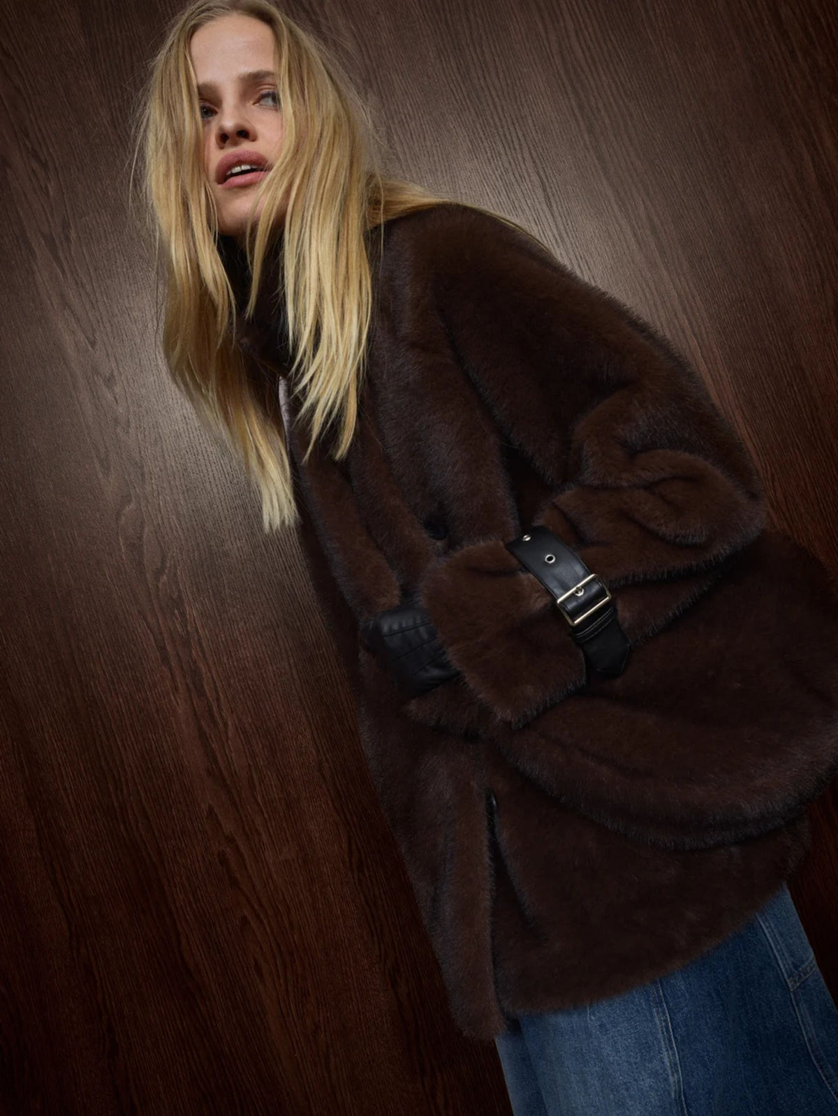 A woman wearing a dark brown plush faux fur coat with a black belt detail, posed against a dark wood-paneled background.