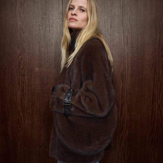 A blonde woman wearing a voluminous dark brown faux-fur jacket and blue jeans, holding a black shoulder bag with a silver buckle, posed against a dark brown wood-paneled wall.