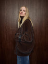 A blonde woman wearing a voluminous dark brown faux-fur jacket and blue jeans, holding a black shoulder bag with a silver buckle, posed against a dark brown wood-paneled wall.